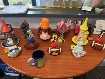 The image features a collection of colorful, hand-painted ceramic figurines arranged on a wooden table. The figurines include a variety of whimsical designs such as gnomes with pointy hats, a teapot with a smiling face, and various themed decorations