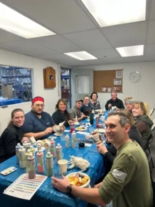 A group of 12 people is seated around a long table covered with a blue tablecloth. They are participating in a painting activity, with various paint bottles and tools in front of them. Some participants are holding ceramic items they’ve painted. The bac