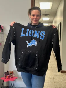 A woman is holding up a black sweatshirt that features the word "LIONS" in large blue letters and an image of a blue lion logo. The sweatshirt has silver studs along the sleeves and pocket. She is smiling and wearing a blue hoodie underneath.