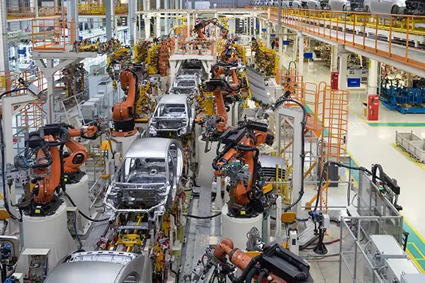 An overhead view of a large automotive manufacturing facility. In the foreground and midground, several partially assembled car frames are positioned on production lines. Robotic arms, mostly orange, are visible on both sides of the car frames, engaged in