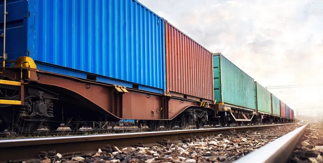 A close-up view of a freight train consisting of多个 colorful shipping containers, including blue, red, and green. The train is positioned on a set of railway tracks, which are lined with gravel. The perspective is low to the ground, focusing on